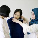 Muslim female adult is trying to fit a new clothes on her younger daughter while the elder daughter is looking on at home.